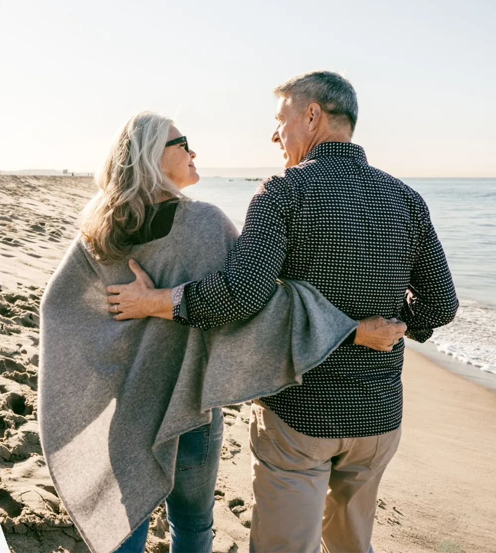 A retired couple walks arm in arm along a beach, smiling at each other.