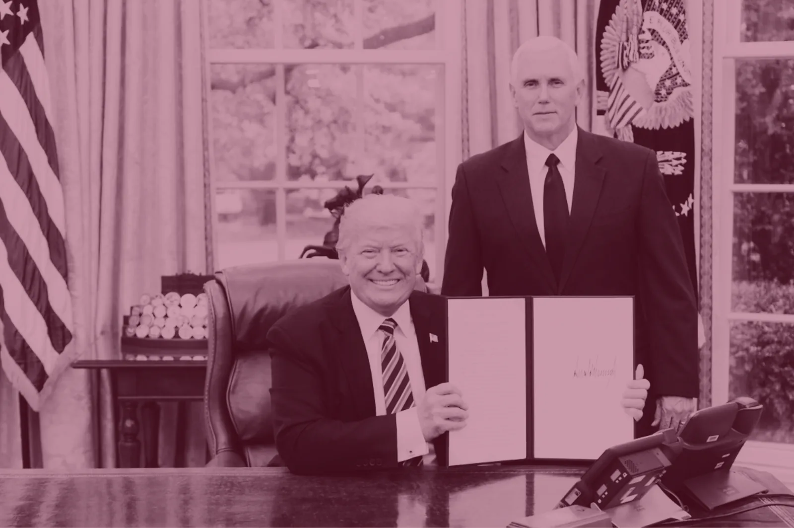 Red-tinted photo of President Trump holding up a signed document at a signing ceremony.